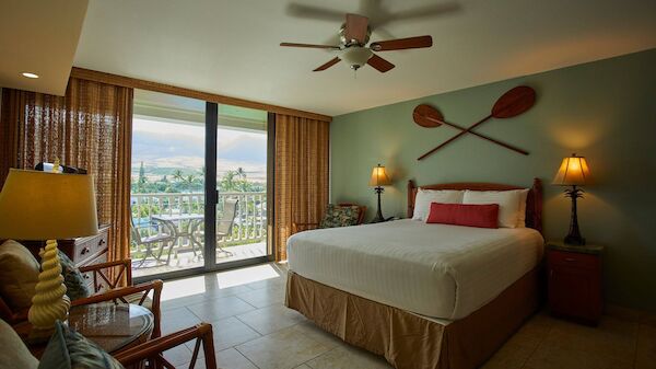 A stylishly decorated hotel room features a double bed, balcony access, and ceiling fan, with decorative paddles on the wall and cozy furnishings.