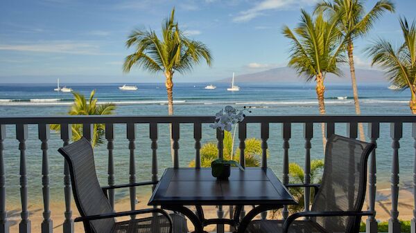 A beautiful ocean view from a balcony, featuring a table with two chairs, palm trees, sailboats, and a calm, blue sea under a clear sky.