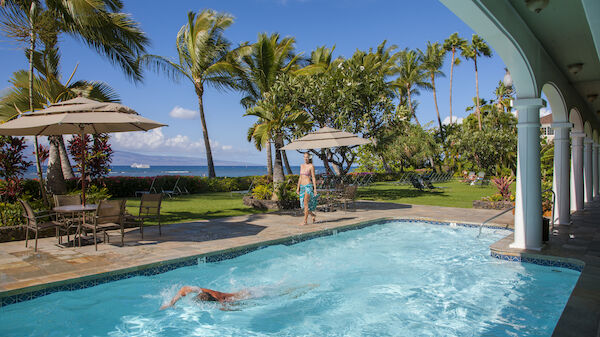 A tropical setting with a swimming pool, palm trees, umbrellas, chairs, a person swimming, and another person walking by the pool under a blue sky.