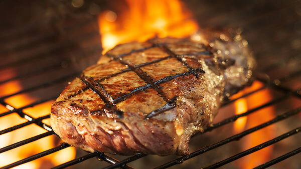 A steak with grill marks is cooking on a grill with flames in the background.