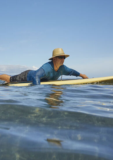 A person wearing a sun hat and blue shirt is lying on a surfboard, paddling on a calm body of water under a clear sky.