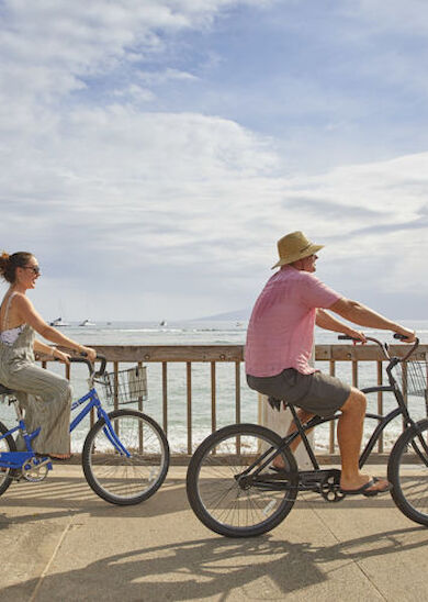 Two people are riding bicycles along a seaside promenade on a bright, sunny day, enjoying the scenic view and pleasant weather.