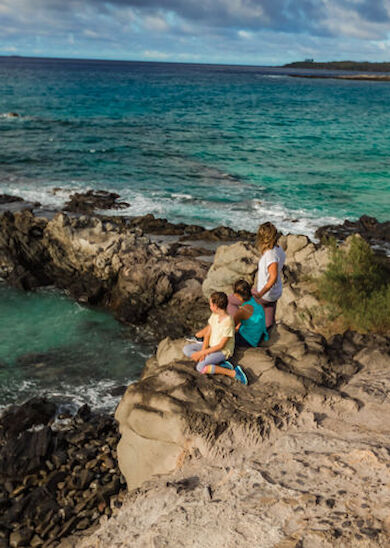 A family of three is standing on rocky cliffs by a turquoise sea, looking out at the horizon under a partly cloudy sky.