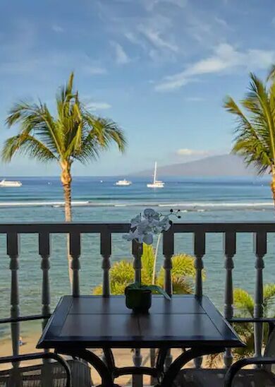 The image shows a balcony with two chairs and a table, overlooking a beach with palm trees, calm waters, and several boats in the distance.