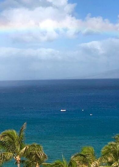 A serene tropical seascape with calm waters, palm trees, and two boats beneath a vibrant rainbow arching across the partly cloudy sky.