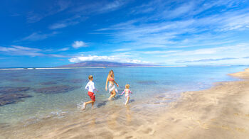 A woman and two children are enjoying a sunny day at the beach, walking along the shore with clear blue skies and calm ocean waters in the background.