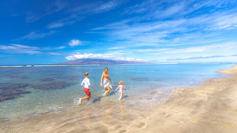 A woman and two children are enjoying a sunny day at the beach, walking along the shore with clear blue skies and calm ocean waters in the background.