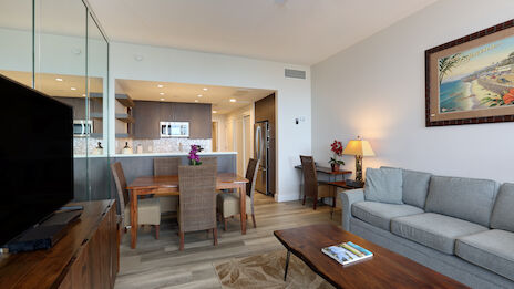 A modern living room with a gray couch, wooden coffee table, TV, dining area, and a kitchen with bar stools is visible. A painting adorns the wall.