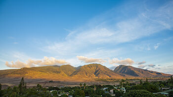 A landscape featuring rolling mountains under a blue sky with scattered clouds, and a green valley with a small collection of buildings.